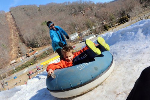 A child in an orange jacket smiles while sitting on a blue snow tube, being pulled up a snowy hill by an adult in a blue jacket.