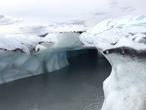 A glacial landscape featuring ice formations and a calm, reflective water surface beneath a cloudy sky.