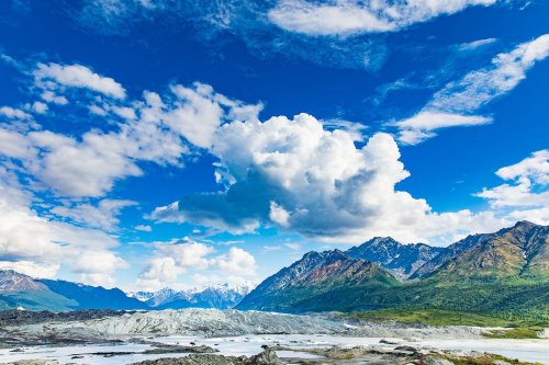 A stunning landscape featuring mountains, a glacier, and a bright blue sky with fluffy clouds.