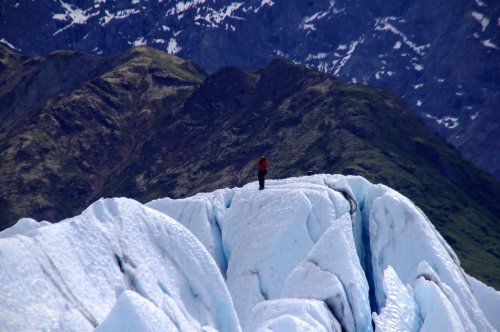 A lone climber stands on a glacier, surrounded by rugged mountains and snow-capped peaks in the background.