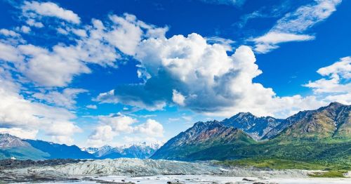 A scenic landscape featuring mountains, a glacier, and a bright blue sky with fluffy clouds.