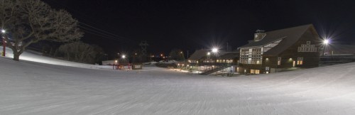 A snowy ski slope at night, illuminated by lights, with a lodge in the background and trees nearby.