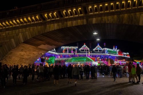 A crowd gathers under a bridge, admiring a brightly lit, festive display of holiday lights on nearby buildings.