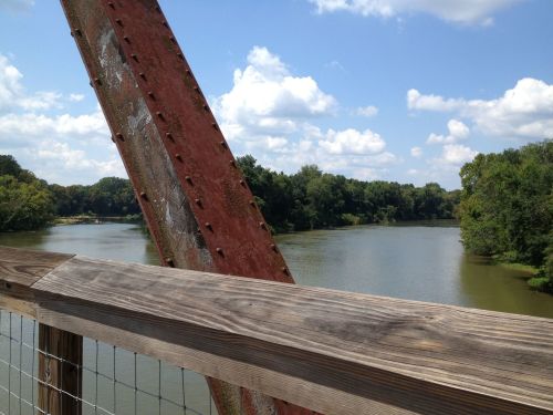 A view of a river surrounded by trees, with a rusty metal bridge support in the foreground under a blue sky.