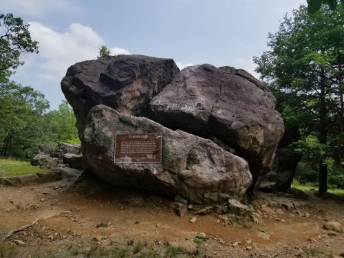 Large boulder with a plaque, surrounded by trees and a clear sky. Ground is rocky and uneven.