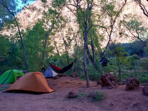 A campsite with two tents and a hammock set among trees, with a rocky landscape and mountains in the background.