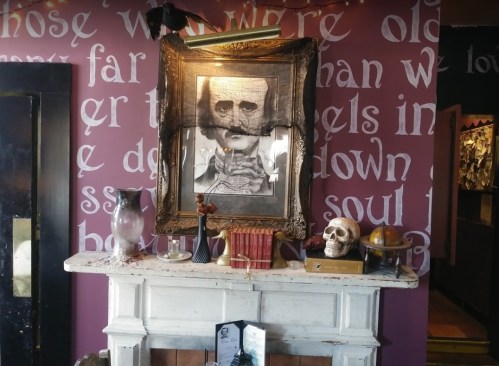 A vintage mantelpiece adorned with a portrait, skull, books, and decorative items against a word-covered wall.