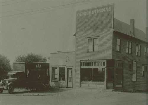 Historic black-and-white photo of a building labeled "George & Thomas" with an old truck parked outside.