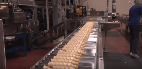 A conveyor belt in a factory filled with ice cream cones, with a worker in the background.