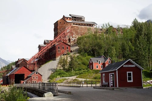 Historic mining buildings in a mountainous area, featuring red structures and a wooden bridge in the foreground.