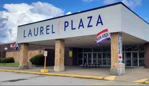 Exterior of Laurel Plaza shopping center with flags and signs, under a blue sky with clouds.