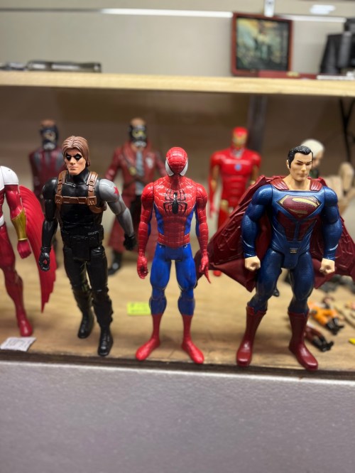 Three action figures stand on a shelf: Spider-Man in red and blue, a character in black, and Superman in blue and red.