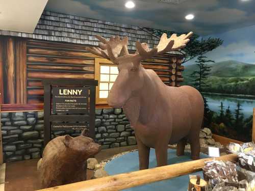 A life-sized moose and bear sculpture inside a rustic log cabin setting, with a scenic mural in the background.