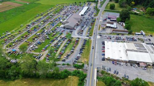 Aerial view of a large outdoor market with numerous cars parked, surrounded by green fields and buildings.
