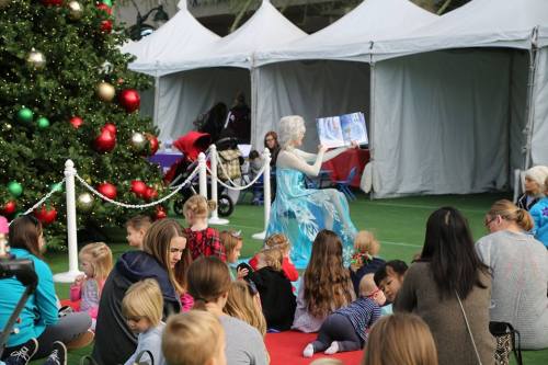A performer dressed as Elsa reads to a group of children seated on the ground near a Christmas tree and tents.