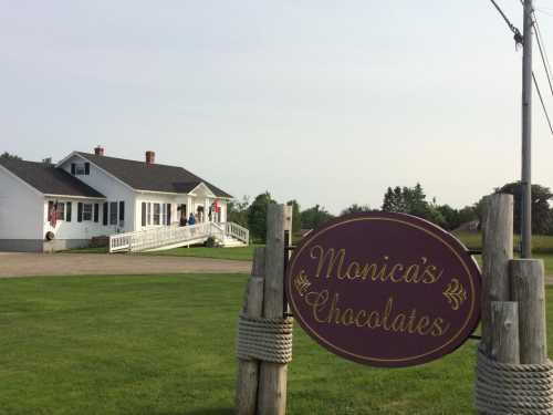 A white house with a porch and American flags, featuring a sign that reads "Monica's Chocolates" in a grassy area.