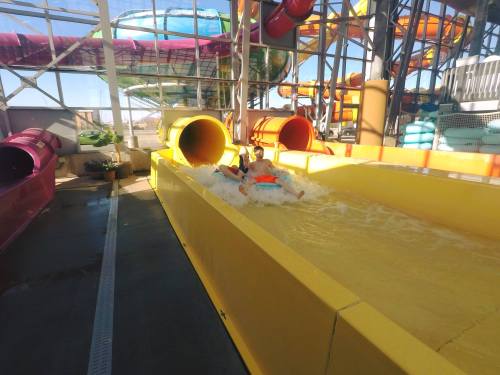 Two children slide down a water slide, splashing into a pool at a water park, surrounded by colorful slides.