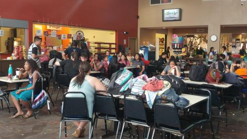 A busy cafeteria with students sitting at tables, chatting, and backpacks scattered around. Bright and lively atmosphere.