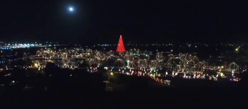 Aerial view of a festive town at night, illuminated with colorful Christmas lights and a large red tree.