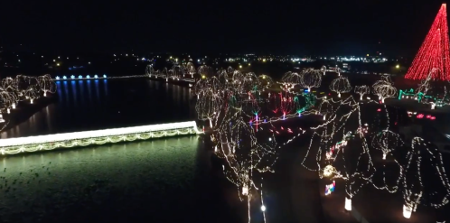 Aerial view of a festive park at night, illuminated with colorful lights and a large red tree by a shimmering lake.