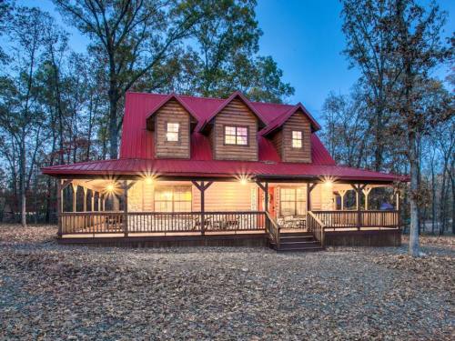 A cozy cabin with a red metal roof, surrounded by trees, illuminated at dusk with a welcoming porch.