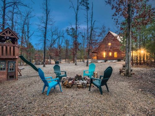 A cozy cabin surrounded by trees, with a fire pit and blue chairs in a gravel clearing at dusk.