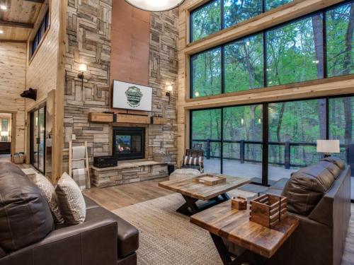 Cozy living room with large windows, stone fireplace, wooden furniture, and a view of the surrounding greenery.