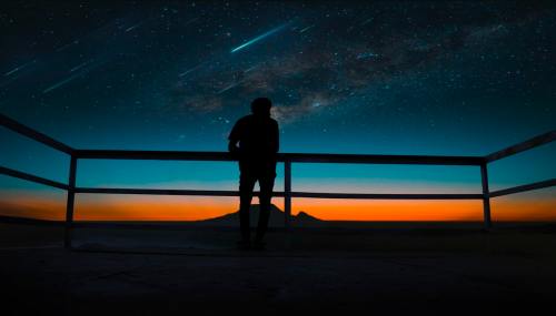 Silhouette of a person gazing at a starry sky with shooting stars and a colorful sunset over distant mountains.