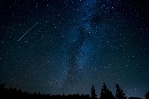 A starry night sky filled with stars and the Milky Way, with a shooting star streaking across the scene.