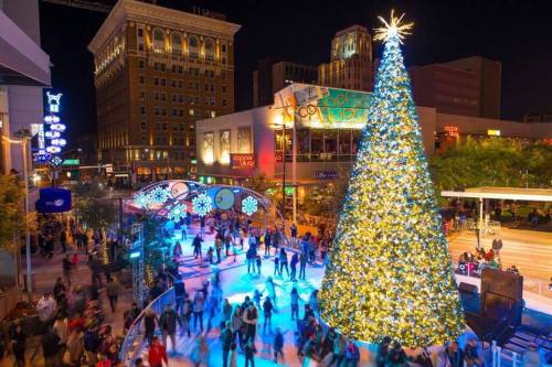 A festive scene with a large Christmas tree, ice skaters, and colorful lights in a bustling urban setting at night.