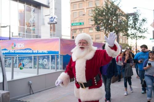 A cheerful Santa Claus waves while walking down a city street, surrounded by people enjoying the festive atmosphere.
