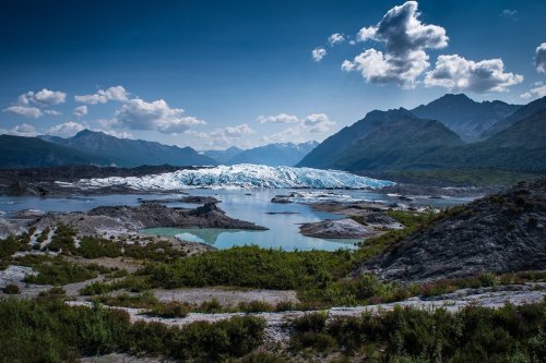 A stunning landscape featuring a glacier surrounded by mountains, a calm lake, and a clear blue sky with clouds.