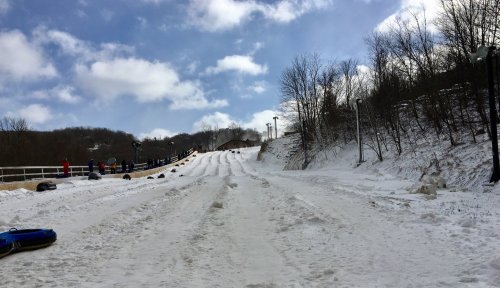 A snowy hill with people sledding, surrounded by trees and a blue sky with clouds.