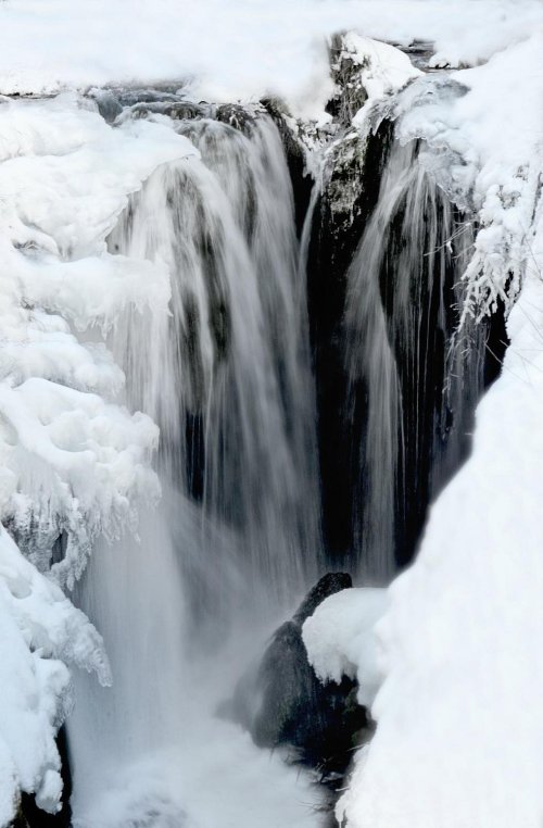 A frozen waterfall cascading over rocks, surrounded by snow and ice, creating a serene winter landscape.