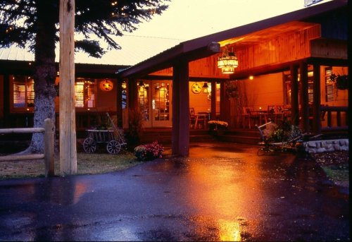 A cozy lodge entrance illuminated at dusk, with wet pavement reflecting warm lights and surrounded by greenery.