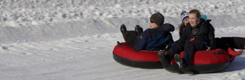 Two people joyfully tubing down a snowy slope on red inflatable tubes. Snowy landscape in the background.