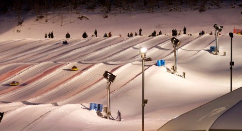 A snowy landscape with multiple tubing lanes, people enjoying the slopes, and floodlights illuminating the scene.