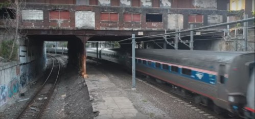 A train speeds through an urban area, passing under a bridge with graffiti on the walls and tracks on either side.