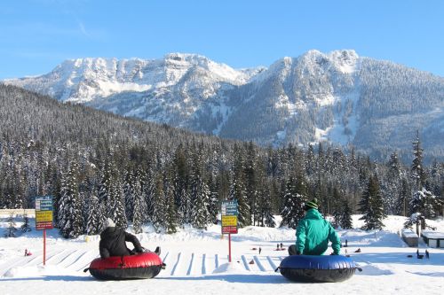 Two people sit on snow tubes, overlooking a snowy landscape with mountains and evergreen trees under a clear blue sky.