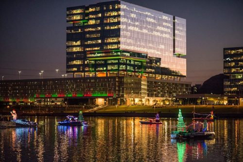 Colorful boats decorated with lights float on a river at night, reflecting a modern building adorned with festive colors.