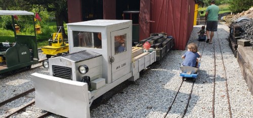 A child rides a small blue cart on tracks beside a model train and a red shed in a scenic outdoor setting.
