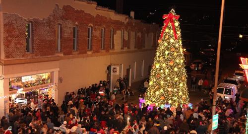 A large, decorated Christmas tree stands in a bustling town square filled with people celebrating the holiday season.
