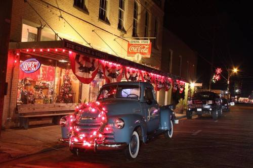 A vintage blue truck adorned with Christmas lights parked outside a festive store decorated for the holidays at night.