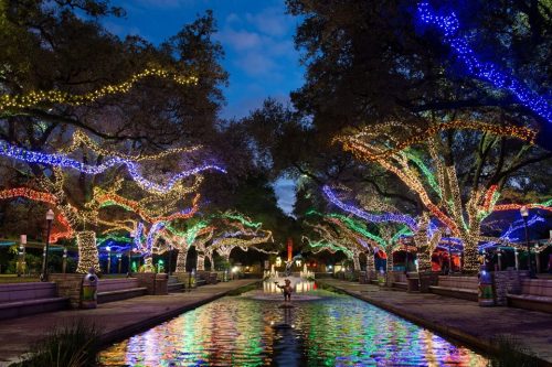 A serene park scene with trees adorned in colorful lights, reflecting in a calm waterway under a twilight sky.