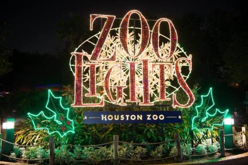 Brightly lit sign reading "Zoo Lights" at Houston Zoo, surrounded by festive decorations and greenery at night.