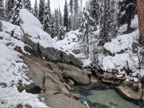 A snowy landscape with rocky terrain and a small, steaming hot spring surrounded by evergreen trees.