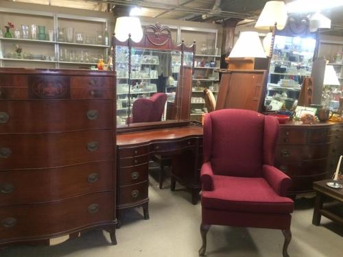 A vintage furniture display featuring a dresser, vanity, and a burgundy armchair in a well-lit shop.