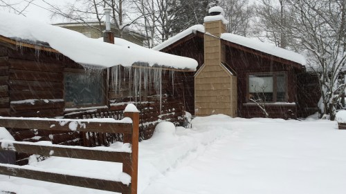 A snowy scene featuring two wooden houses with snow-covered roofs and icicles, surrounded by a white landscape.