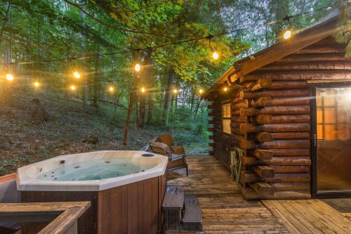 A cozy cabin with a hot tub on a wooden deck, surrounded by trees and illuminated by string lights.