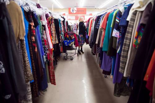 A woman stands between rows of colorful clothing in a thrift store, pushing a shopping cart filled with clothes.